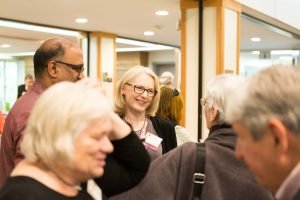 Blonde woman wearing glasses stands in center of group of people.