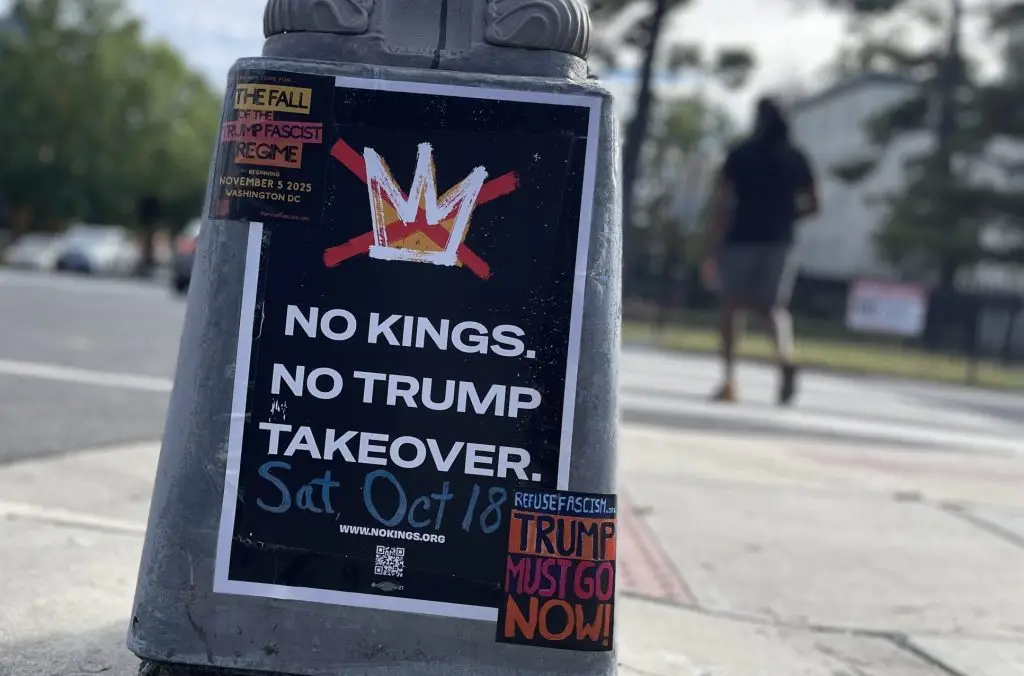 Street scene with a gray pole in the center foreground showing a poster with black background and large white lettering, "No Kings. No Trump Takeover. Sat, Oct 18."