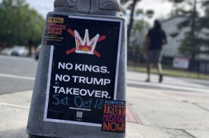 Street scene with a gray pole in the center foreground showing a poster with black background and large white lettering, "No Kings. No Trump Takeover. Sat, Oct 18."