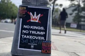 Street scene with a gray pole in the center foreground showing a poster with black background and large white lettering, "No Kings. No Trump Takeover. Sat, Oct 18."